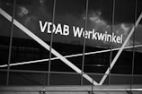 The image depicts a large window with the text 'VDAB Werkwinkel' prominently displayed on it. The glass has diagonal white lines across it and the background reflects a cloudy sky and faint structures.