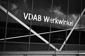 The image depicts a large window with the text 'VDAB Werkwinkel' prominently displayed on it. The glass has diagonal white lines across it and the background reflects a cloudy sky and faint structures.