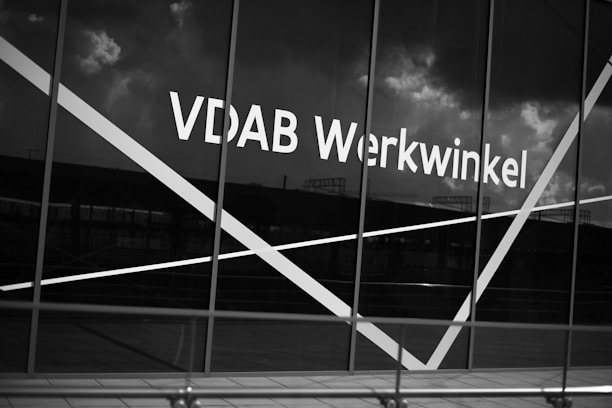 The image depicts a large window with the text 'VDAB Werkwinkel' prominently displayed on it. The glass has diagonal white lines across it and the background reflects a cloudy sky and faint structures.