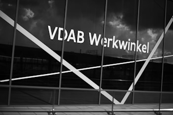 The image depicts a large window with the text 'VDAB Werkwinkel' prominently displayed on it. The glass has diagonal white lines across it and the background reflects a cloudy sky and faint structures.