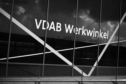 The image depicts a large window with the text 'VDAB Werkwinkel' prominently displayed on it. The glass has diagonal white lines across it and the background reflects a cloudy sky and faint structures.