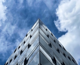 A modern glass building rises toward the sky, reflecting the fluffy white clouds and deep blue sky in its mirrored surface. The architecture features clean lines and geometric shapes, creating a sleek and contemporary appearance.