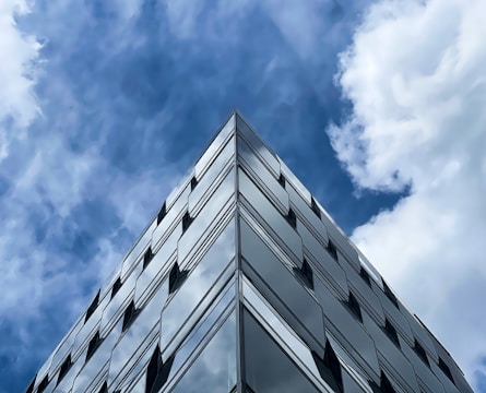 A modern glass building rises toward the sky, reflecting the fluffy white clouds and deep blue sky in its mirrored surface. The architecture features clean lines and geometric shapes, creating a sleek and contemporary appearance.