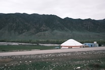 A wide plain with a large white yurt and adjacent structures set against a backdrop of towering green mountains. In the foreground, there is an asphalt road and patches of green grass with scattered stones. The sky is overcast and the landscape is vast and open.