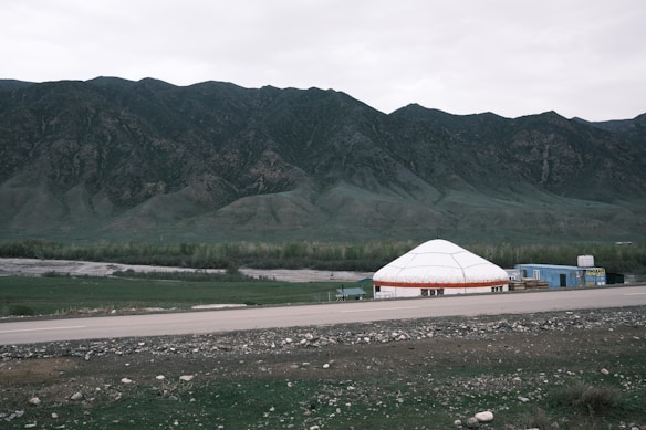 A wide plain with a large white yurt and adjacent structures set against a backdrop of towering green mountains. In the foreground, there is an asphalt road and patches of green grass with scattered stones. The sky is overcast and the landscape is vast and open.