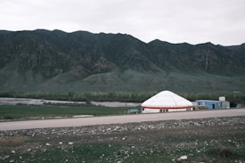A wide plain with a large white yurt and adjacent structures set against a backdrop of towering green mountains. In the foreground, there is an asphalt road and patches of green grass with scattered stones. The sky is overcast and the landscape is vast and open.
