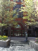 A red-brick building with multiple floors features a prominent arched entrance labeled 'Tent City'. The entrance is flanked by two metal railings and surrounded by blooming trees and flower beds with vibrant yellow and purple flowers. The sunlight casts shadows on the pavement, creating a welcoming and serene atmosphere.