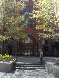 A red-brick building with multiple floors features a prominent arched entrance labeled 'Tent City'. The entrance is flanked by two metal railings and surrounded by blooming trees and flower beds with vibrant yellow and purple flowers. The sunlight casts shadows on the pavement, creating a welcoming and serene atmosphere.