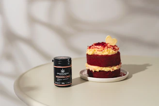 A small, layered red velvet cake with cream cheese frosting sits on a white plate. Beside it, a black jar labeled 'Crystalweed Wedding Cake' with a cannabis leaf logo rests on a light-colored table. The cake is topped with a light yellow heart-shaped decoration and red crumbs.