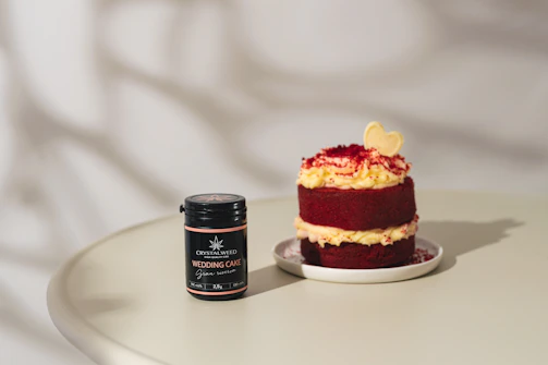 A small, layered red velvet cake with cream cheese frosting sits on a white plate. Beside it, a black jar labeled 'Crystalweed Wedding Cake' with a cannabis leaf logo rests on a light-colored table. The cake is topped with a light yellow heart-shaped decoration and red crumbs.