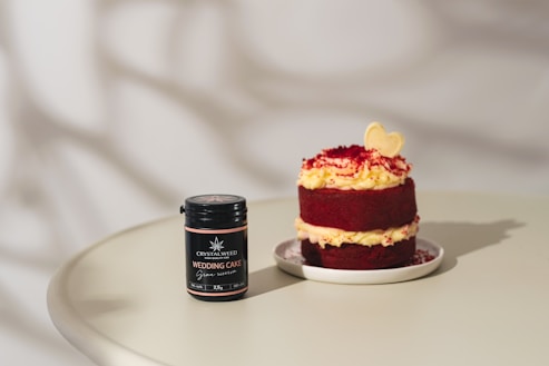 A small, layered red velvet cake with cream cheese frosting sits on a white plate. Beside it, a black jar labeled 'Crystalweed Wedding Cake' with a cannabis leaf logo rests on a light-colored table. The cake is topped with a light yellow heart-shaped decoration and red crumbs.