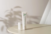 A tube of organic cream labeled 'beauty' is placed upright on a round surface. Soft shadows and ambient lighting create a serene atmosphere. The background is minimal with abstract shadow patterns, suggesting a natural theme.