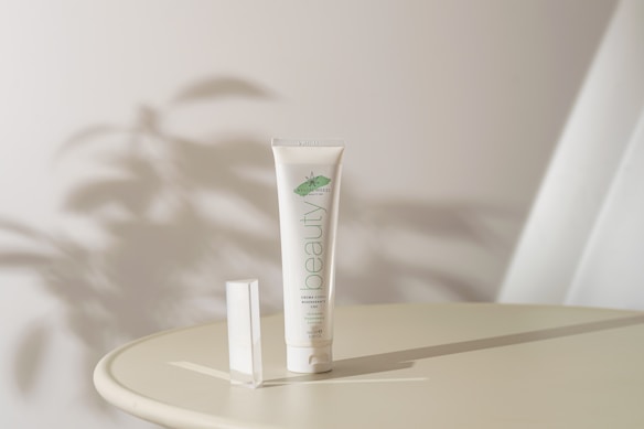 A tube of organic cream labeled 'beauty' is placed upright on a round surface. Soft shadows and ambient lighting create a serene atmosphere. The background is minimal with abstract shadow patterns, suggesting a natural theme.