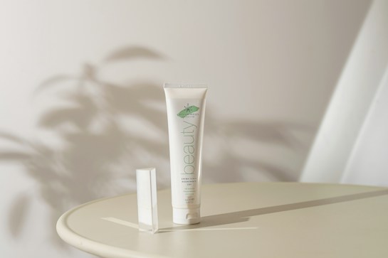 A tube of organic cream labeled 'beauty' is placed upright on a round surface. Soft shadows and ambient lighting create a serene atmosphere. The background is minimal with abstract shadow patterns, suggesting a natural theme.