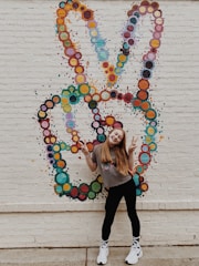 A young person with long hair is posing happily in front of a colorful mural. The mural features a peace sign created from clusters of painted circles in various hues. The person is making a peace sign with both hands, dressed in casual clothing including a gray t-shirt, black leggings, and white sneakers. The overall setting is an urban brick wall, adding to the vibrant and lively atmosphere.