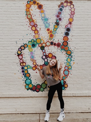 A young person with long hair is posing happily in front of a colorful mural. The mural features a peace sign created from clusters of painted circles in various hues. The person is making a peace sign with both hands, dressed in casual clothing including a gray t-shirt, black leggings, and white sneakers. The overall setting is an urban brick wall, adding to the vibrant and lively atmosphere.
