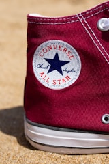 A close-up of a maroon Converse Chuck Taylor All Star sneaker resting on a sandy surface. The classic white circular logo with a blue star is prominently visible on the side of the shoe.