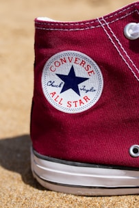 A close-up of a maroon Converse Chuck Taylor All Star sneaker resting on a sandy surface. The classic white circular logo with a blue star is prominently visible on the side of the shoe.