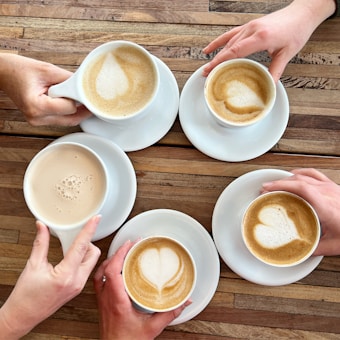 Five cups of coffee arranged in a circle on a wooden table, each cup showing latte art in the shape of a heart. The hands of different individuals are holding the cups, suggesting a shared coffee moment.