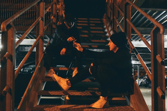 Two individuals are seated on a metal staircase under dim lighting. One person is sitting on a step while the other is crouched higher up, and they appear to be exchanging an item. The scene is set in an industrial-like environment with visible metal beams and railings.