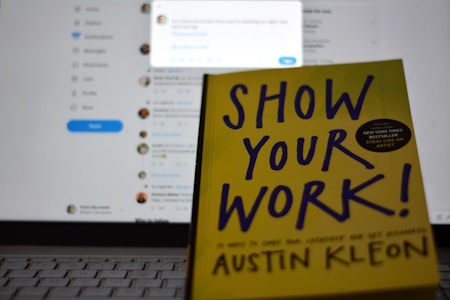 A book titled 'Show Your Work!' by Austin Kleon is propped open in front of a laptop displaying a blurred social media interface. The book cover is bright yellow with bold, dark blue text.
