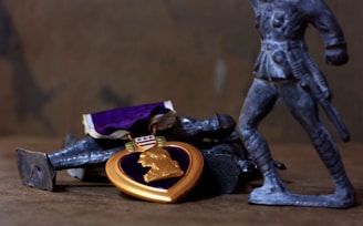 A Purple Heart medal rests on a surface next to metallic toy soldiers. The medal has a purple background with gold detailing, including a gold ribbon and a central portrait. The toy soldiers are gray and positioned in various stances, with one standing prominently in the foreground.