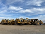 Several large, yellow construction vehicles, likely road scrapers or earth movers, are parked on a dirt surface under a partly cloudy sky. The vehicles are arranged in a row, showcasing their massive wheels and equipment.