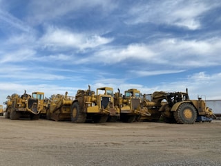Several large, yellow construction vehicles, likely road scrapers or earth movers, are parked on a dirt surface under a partly cloudy sky. The vehicles are arranged in a row, showcasing their massive wheels and equipment.