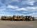 Several large, yellow construction vehicles, likely road scrapers or earth movers, are parked on a dirt surface under a partly cloudy sky. The vehicles are arranged in a row, showcasing their massive wheels and equipment.