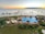 A luxurious beachfront resort featuring a large, elegantly designed outdoor swimming pool surrounded by palm trees and gardens. Lounge chairs with umbrellas are arranged around the pool area. The ocean extends into the horizon with an island visible in the distance under a sunset sky.