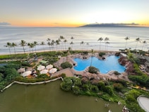 A luxurious beachfront resort featuring a large, elegantly designed outdoor swimming pool surrounded by palm trees and gardens. Lounge chairs with umbrellas are arranged around the pool area. The ocean extends into the horizon with an island visible in the distance under a sunset sky.