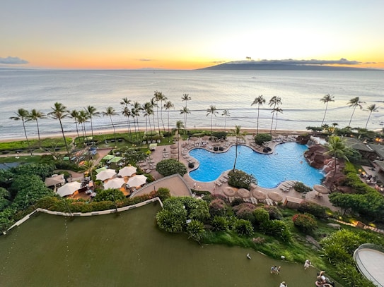 A luxurious beachfront resort featuring a large, elegantly designed outdoor swimming pool surrounded by palm trees and gardens. Lounge chairs with umbrellas are arranged around the pool area. The ocean extends into the horizon with an island visible in the distance under a sunset sky.