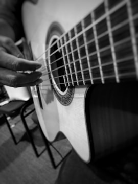 A close-up view of a person's hand playing a classical acoustic guitar. The image is in black and white, emphasizing the textures of the guitar strings and the intricate design around the sound hole. The background appears to be a room with chairs.