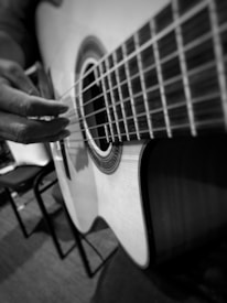 A close-up view of a person's hand playing a classical acoustic guitar. The image is in black and white, emphasizing the textures of the guitar strings and the intricate design around the sound hole. The background appears to be a room with chairs.