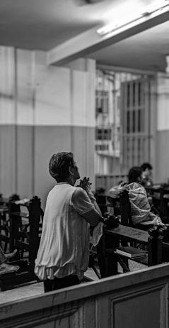 An elderly person is seen in a grayscale image, kneeling on a pew in what appears to be a place of worship or a chapel. The individual is praying with clasped hands. The space is dimly lit, and there are other people seated at a distance, some covered with blankets or cloaks. The architecture is simple, with paneled walls and a barred window.