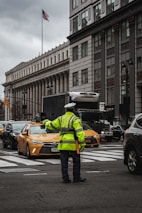 A traffic officer is directing vehicles at a busy intersection in an urban setting. Surrounding the scene are large buildings with classical architecture, including columns and detailed facades. The American flag is visible on a flagpole, and there are yellow taxis and various other vehicles on the road.