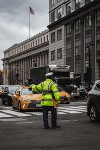A traffic officer is directing vehicles at a busy intersection in an urban setting. Surrounding the scene are large buildings with classical architecture, including columns and detailed facades. The American flag is visible on a flagpole, and there are yellow taxis and various other vehicles on the road.