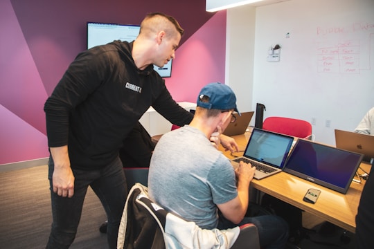 A group of men in a modern office setting. Two individuals are focused on a laptop screen, collaborating over something displayed. The room has a casual atmosphere with a modern layout, featuring multiple laptops on wooden tables and a digital whiteboard mounted on the walled section painted pink and white.
