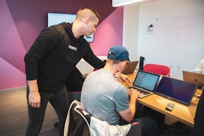 A group of men in a modern office setting. Two individuals are focused on a laptop screen, collaborating over something displayed. The room has a casual atmosphere with a modern layout, featuring multiple laptops on wooden tables and a digital whiteboard mounted on the walled section painted pink and white.
