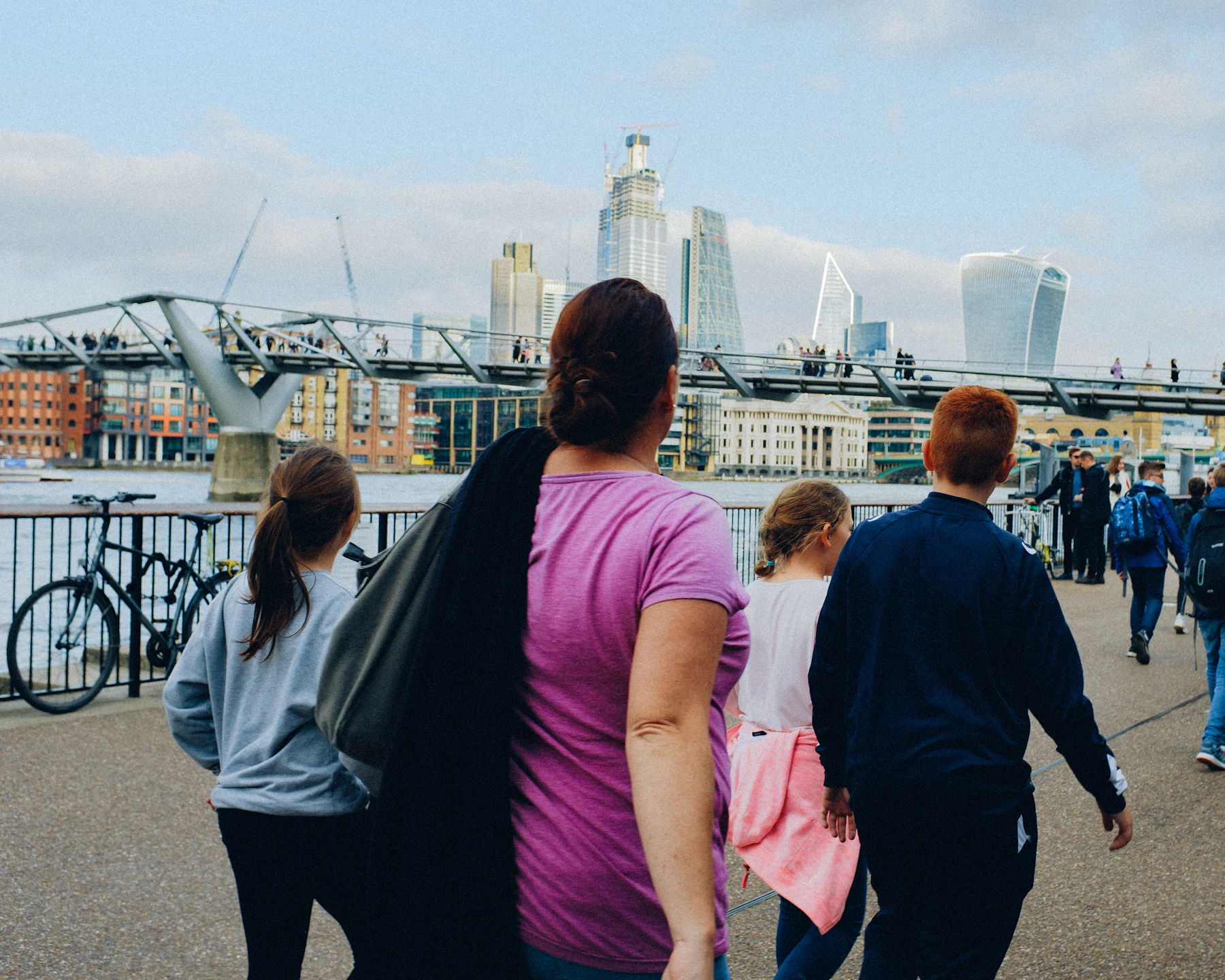 Photograph of An adult and three children walk beside the Thames with London’s skyline and a footbridge ahead.