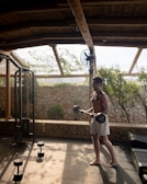 A man is working out in an open-air gym, lifting weights in a space with a stone wall and wooden roof. The gym is surrounded by natural greenery, and there's a fan attached to a wooden beam. Sunlight filters through the wooden slats, casting shadows on the floor.
