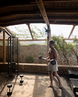 A man is working out in an open-air gym, lifting weights in a space with a stone wall and wooden roof. The gym is surrounded by natural greenery, and there's a fan attached to a wooden beam. Sunlight filters through the wooden slats, casting shadows on the floor.