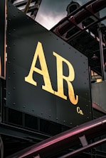 A close-up view of a metal signboard with large, gold-colored letters 'AR Co.' against a dark background. The sign is part of an industrial structure with visible metal beams and pipes surrounding it.