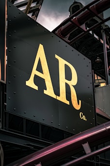 A close-up view of a metal signboard with large, gold-colored letters 'AR Co.' against a dark background. The sign is part of an industrial structure with visible metal beams and pipes surrounding it.