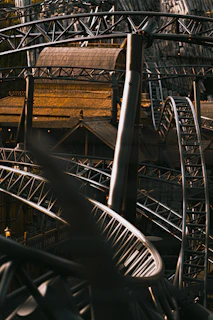 Complex steel roller coaster tracks intertwine, creating dynamic loops and curves against a backdrop of wooden and metal structures. The lighting casts a warm glow, accentuating the intricate design and architecture.