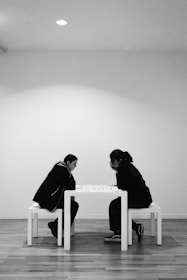 Two individuals are deeply focused on a game of chess. They sit across from each other at a simple white table, positioned against a minimalistic background. The setting appears to be a bright, minimally furnished room with white walls and wooden flooring, creating a calm and contemplative atmosphere.