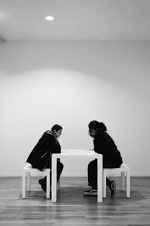 Two individuals are deeply focused on a game of chess. They sit across from each other at a simple white table, positioned against a minimalistic background. The setting appears to be a bright, minimally furnished room with white walls and wooden flooring, creating a calm and contemplative atmosphere.