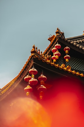 An ornate rooftop with intricate golden details and decorative elements displays traditional architecture. Several red lanterns hang elegantly from the building, contrasting against the clear blue sky. A warm, hazy orange hue adds a soft, atmospheric touch to the lower part of the image.