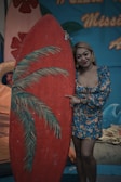 A woman wearing a blue floral dress stands next to a large red surfboard with painted palm leaves. The background features a tropical theme with blue and orange decorative elements, including text in orange script.