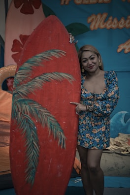 A woman wearing a blue floral dress stands next to a large red surfboard with painted palm leaves. The background features a tropical theme with blue and orange decorative elements, including text in orange script.
